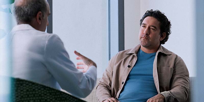 Photo of an adult male sitting on sofa consulting with another adult male in the foreground