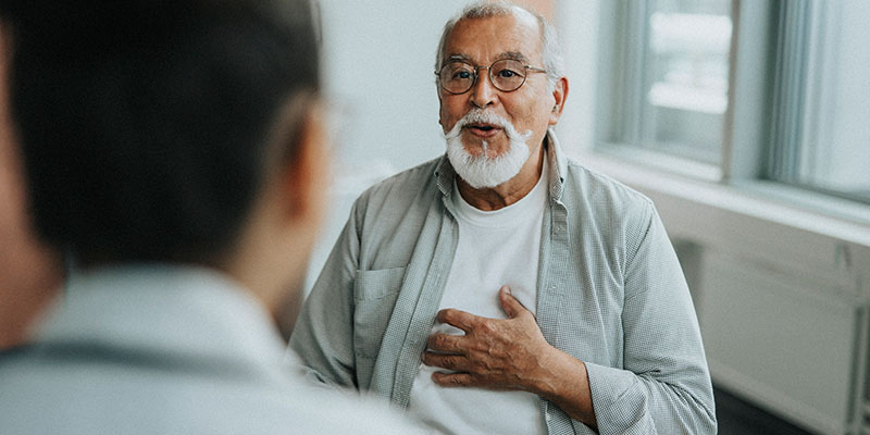 Photo of an adult male seated in the background with his hand on his chest as he is consulting with a clinician in the foreground