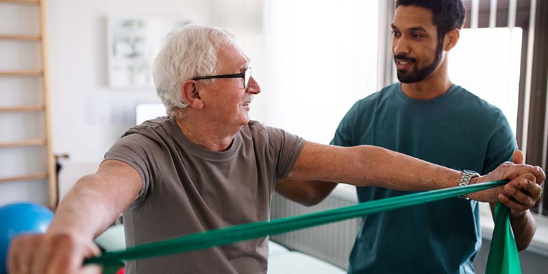Photo of an older adult male using a resistance band while being coached by a physical therapist