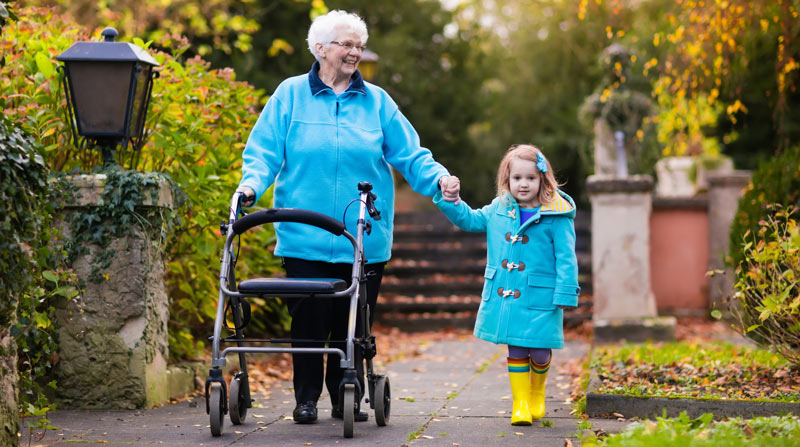 An elderly woman walking outdoors in the fall, with a walker and holding hands with a child.